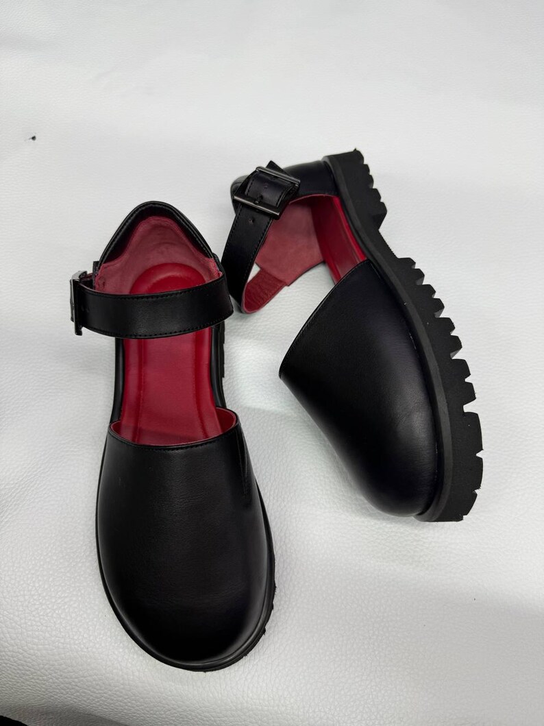 May include: Pair of black Mary Jane style shoes with a red interior. The shoes have a thick, lug sole and a buckle closure on the ankle strap. The shoes are made of a smooth material.