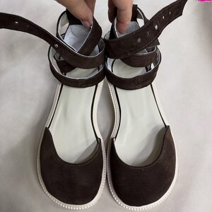 May include: A pair of brown suede sandals with white soles and straps. The sandals have a flat design with a rounded toe and ankle straps. The straps are adjustable and feature a buckle closure.