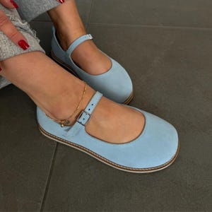 May include: Pale blue Mary Jane style flats with a strap and buckle fastening. The shoes have a rounded toe and a light brown sole. The shoes are made of a suede-like material.