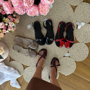 May include: A collection of women's sandals in various colors, including black, red, and beige. The sandals have a minimalist design with straps and buckles. The image also features a display of pink roses and decorative woven placemats, creating a stylish presentation.