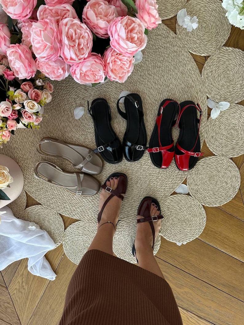 May include: A collection of women's sandals in various colors, including black, red, and beige. The sandals have a minimalist design with straps and buckles. The image also features a display of pink roses and decorative woven placemats, creating a stylish presentation.