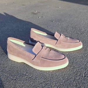 May include: A pair of mauve suede loafers featuring a classic moccasin-style toe, a decorative strap across the vamp, and a thick, solid white rubber sole. The interior lining is a light, pale green color, providing a contrast to the exterior.