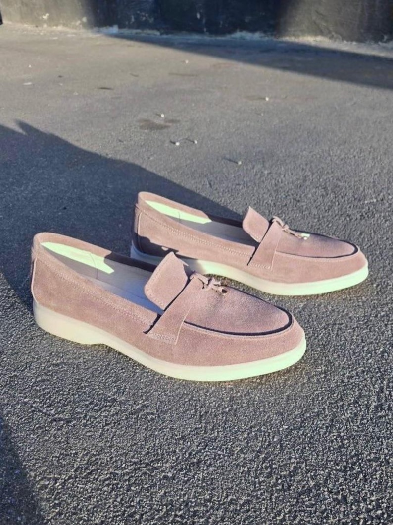 May include: A pair of mauve suede loafers featuring a classic moccasin-style toe, a decorative strap across the vamp, and a thick, solid white rubber sole. The interior lining is a light, pale green color, providing a contrast to the exterior.