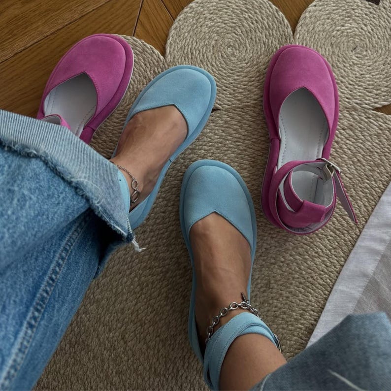 May include: A pair of light blue suede flats and a pair of magenta suede sandals with ankle straps. The shoes are displayed on a woven mat, with a person wearing the blue flats. The shoes have a minimalist design.