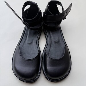 May include: A pair of black leather sandals with a flat sole. The sandals have an ankle strap and a strap that goes around the ankle. The sandals are made of a smooth, shiny leather material.