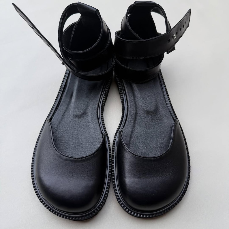 May include: A pair of black leather sandals with a flat sole. The sandals have an ankle strap and a strap that goes around the ankle. The sandals are made of a smooth, shiny leather material.