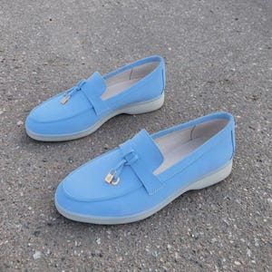 May include: A pair of light blue loafers featuring a smooth matte finish, a rounded toe, and a decorative strap across the vamp. Each shoe is accented with a small gold-toned metal padlock charm and sits on a thick, light gray rubber sole.