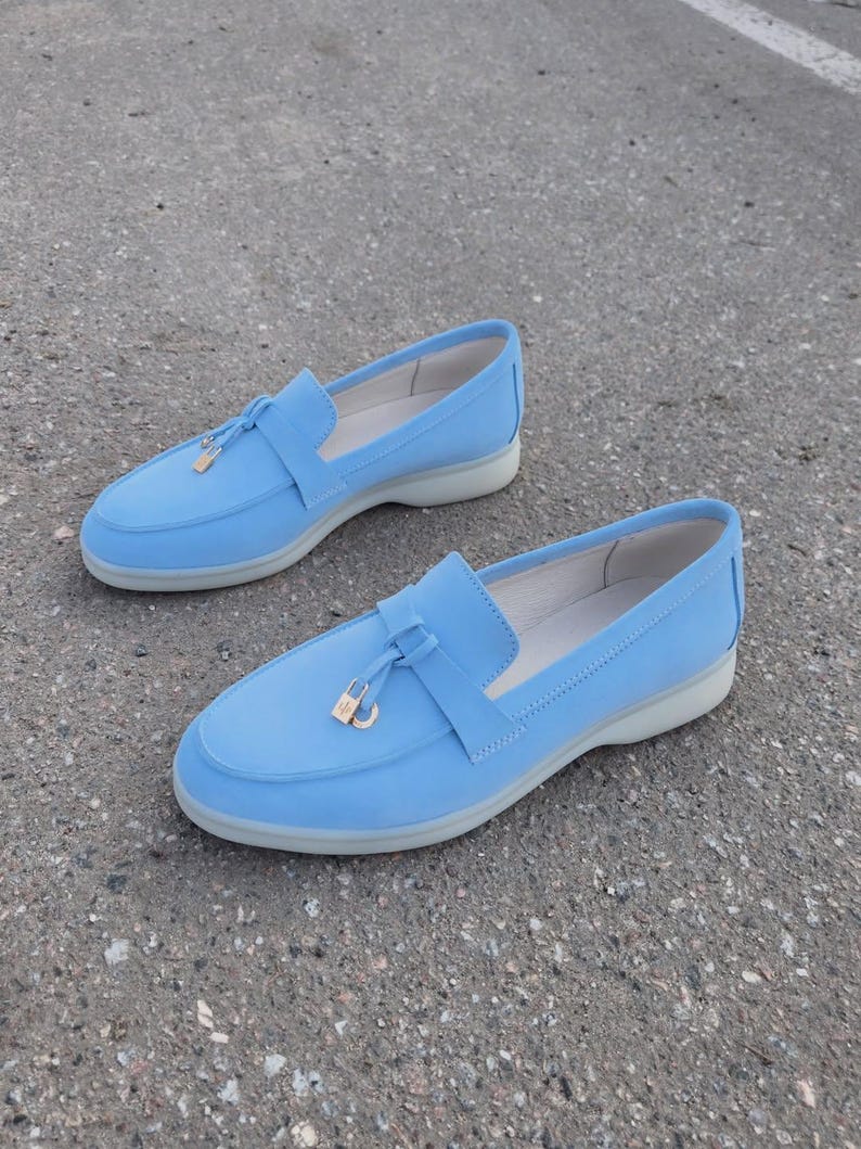 May include: A pair of light blue loafers featuring a smooth matte finish, a rounded toe, and a decorative strap across the vamp. Each shoe is accented with a small gold-toned metal padlock charm and sits on a thick, light gray rubber sole.