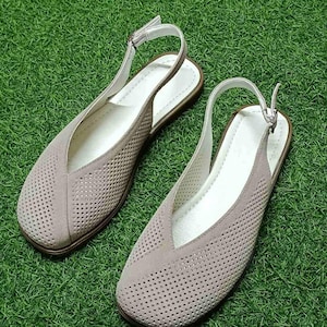 May include: A pair of taupe-colored slingback flats with a perforated design. The shoes have a V-shaped vamp, white insoles, and adjustable ankle straps. The soles are brown, and the shoes are set against a green background.