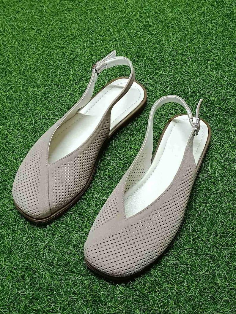 May include: A pair of taupe-colored slingback flats with a perforated design. The shoes have a V-shaped vamp, white insoles, and adjustable ankle straps. The soles are brown, and the shoes are set against a green background.