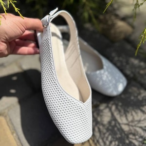 May include: White perforated leather flats with a slingback strap and a small buckle. The shoes have a square toe and a neutral-colored insole. A second pair of silver flats is slightly out of focus in the background.