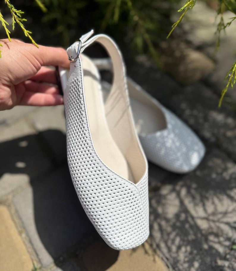 May include: White perforated leather flats with a slingback strap and a small buckle. The shoes have a square toe and a neutral-colored insole. A second pair of silver flats is slightly out of focus in the background.