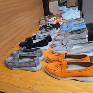 May include: A collection of suede loafers arranged in rows on a wooden surface. The shoes feature a tassel detail on the front, a rounded toe, and a thick white rubber sole. Available colors include bright orange, light gray, black, and light blue.