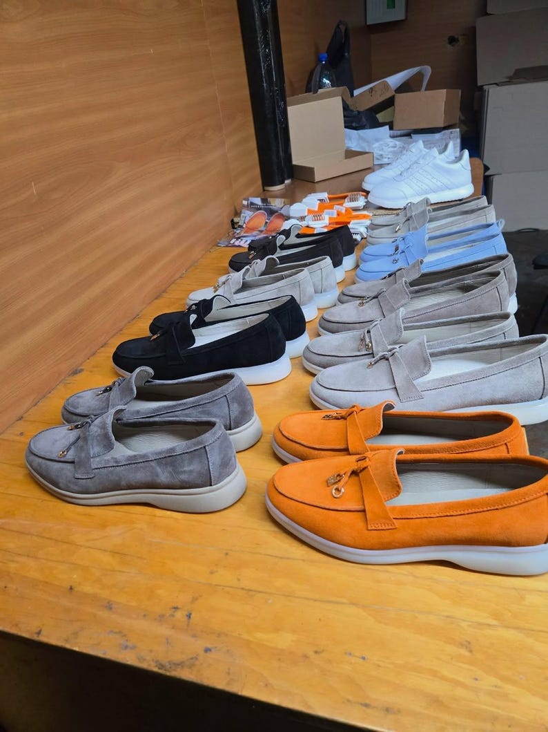 May include: A collection of suede loafers arranged in rows on a wooden surface. The shoes feature a tassel detail on the front, a rounded toe, and a thick white rubber sole. Available colors include bright orange, light gray, black, and light blue.