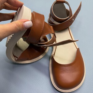 May include: Brown leather sandals with a rounded toe and ankle strap. The sandals have a thick, off-white sole and a buckle closure. The design features a strap that wraps around the ankle.