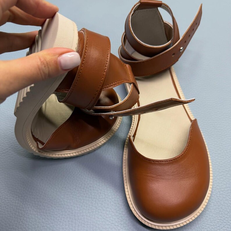 May include: Brown leather sandals with a rounded toe and ankle strap. The sandals have a thick, off-white sole and a buckle closure. The design features a strap that wraps around the ankle.