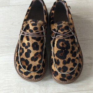 Pu&ograve; includere: Un paio di scarpe slip-on con stampa leopardata. Le scarpe hanno una suola marrone e un motivo maculato marrone e nero. Le scarpe hanno lacci marroni e un bordo cucito intorno alla suola.