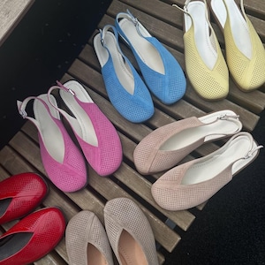 May include: A collection of colorful, perforated leather flats in various shades, including red, pink, blue, yellow, and beige. Each pair features a slingback strap with a silver buckle. The shoes are arranged on a wooden surface.