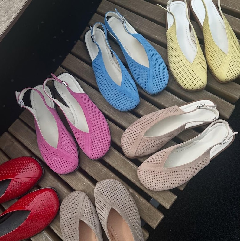 May include: A collection of colorful, perforated leather flats in various shades, including red, pink, blue, yellow, and beige. Each pair features a slingback strap with a silver buckle. The shoes are arranged on a wooden surface.