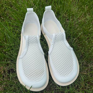 May include: A pair of white slip-on shoes with a mesh vamp and a rounded toe. The shoes have a light beige sole and are set against a backdrop of green grass. The shoes appear to be made of leather or a similar material.