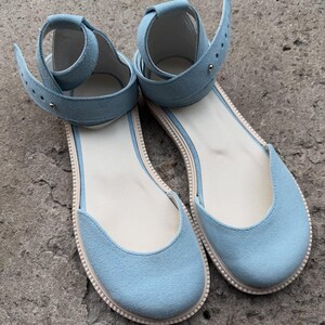 May include: A pair of light blue suede sandals with ankle straps. The sandals have a flat sole with a cream-colored insole and a light beige trim. The straps are adjustable with a buckle closure. The shoes are designed for casual wear.