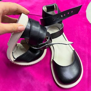 May include: Black leather sandals with a white sole and multiple straps. The sandals have a rounded toe and an ankle strap with a buckle closure. The sole appears to be approximately 1 inch thick.
