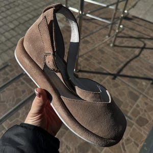 May include: Taupe suede sandal with a rounded toe and ankle strap. The sandal features a buckle closure and a white sole. The shoe has a wedge heel and a strap that goes around the ankle.