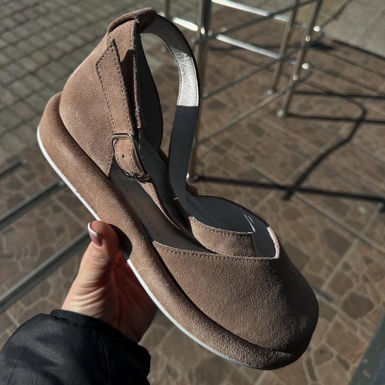 May include: Taupe suede sandal with a rounded toe and ankle strap. The sandal features a buckle closure and a white sole. The shoe has a wedge heel and a strap that goes around the ankle.