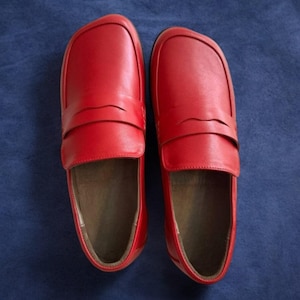May include: A pair of bright red leather loafers. The shoes have a classic design with a rounded toe and a strap detail across the top. The interior of the shoes is a light brown color. The loafers are displayed on a blue surface.
