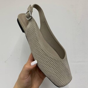 May include: A beige, perforated leather slingback flat shoe with a square toe. The shoe features a buckle closure and a small heel. The shoe is held by a hand with a white painted nail.