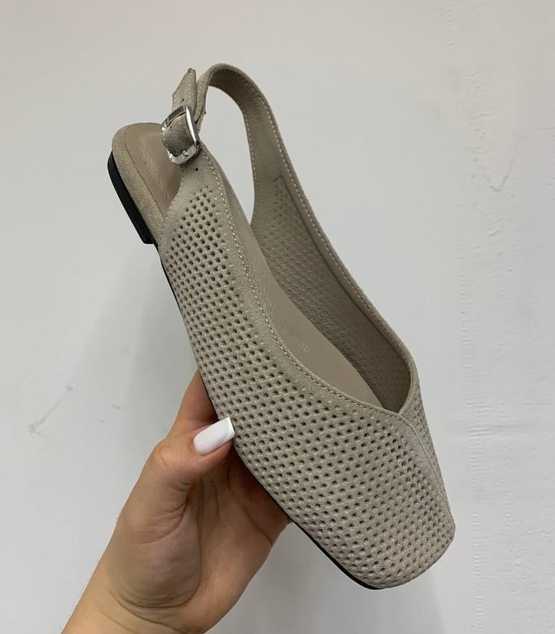 May include: A beige, perforated leather slingback flat shoe with a square toe. The shoe features a buckle closure and a small heel. The shoe is held by a hand with a white painted nail.