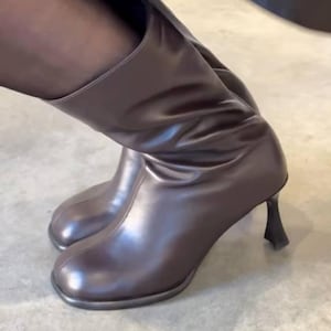 Op de afbeelding: Bruine leren kniehoge laarzen met een ronde neus en een unieke, sculpturale hak. De laarzen hebben een glanzende afwerking. De schoenen zijn een modieus item. De laarzen zijn ongeveer 30 cm hoog.
