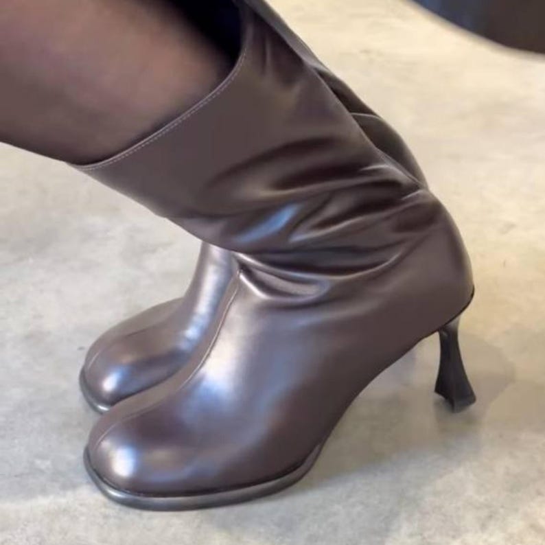 Op de afbeelding: Bruine leren kniehoge laarzen met een ronde neus en een unieke, sculpturale hak. De laarzen hebben een glanzende afwerking. De schoenen zijn een modieus item. De laarzen zijn ongeveer 30 cm hoog.