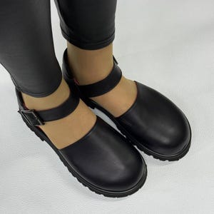 May include: Black leather Mary Jane style shoes with a thick sole and a buckle closure on the ankle strap. The shoes have a rounded toe and a textured sole. The shoes are worn with black leggings.