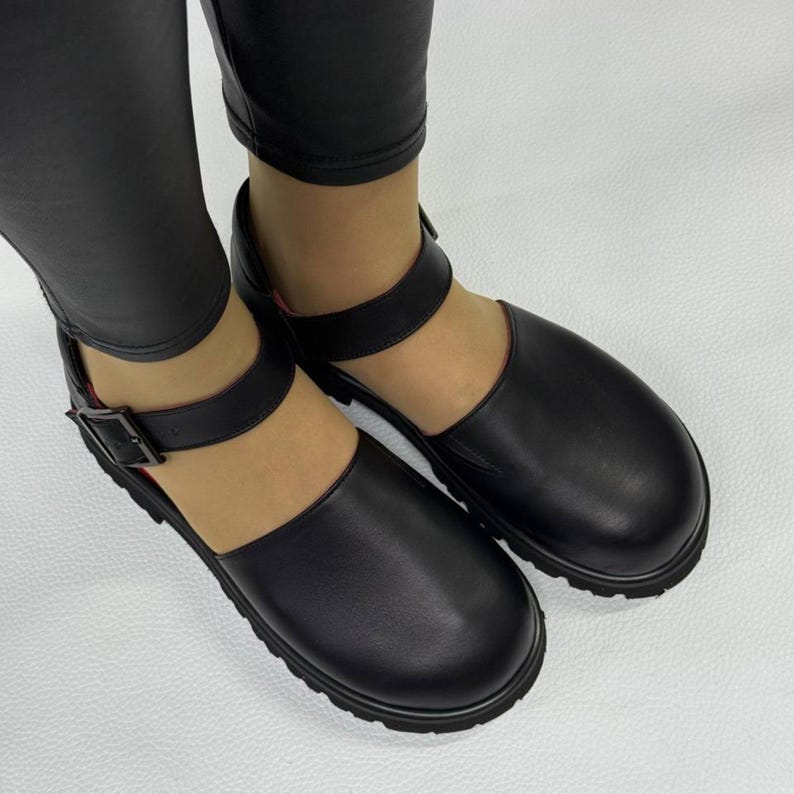 May include: Black leather Mary Jane style shoes with a thick sole and a buckle closure on the ankle strap. The shoes have a rounded toe and a textured sole. The shoes are worn with black leggings.