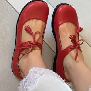 May include: A pair of red Mary Jane style shoes with black soles. The shoes have a strap across the top of the foot with a tassel and a gold chain ankle bracelet. The shoes are made of a smooth material.