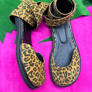 May include: A pair of leopard print sandals with black soles and straps. The sandals have a flat sole and a wrap-around ankle strap. The leopard print features brown and black spots on a tan background. The sandals are displayed on a pink surface.