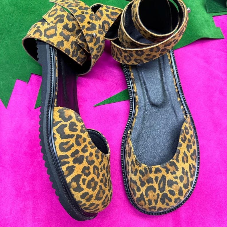 May include: A pair of leopard print sandals with black soles and straps. The sandals have a flat sole and a wrap-around ankle strap. The leopard print features brown and black spots on a tan background. The sandals are displayed on a pink surface.