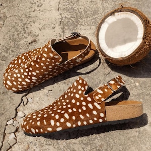 Puede incluir: Un par de zuecos con estampado animal de manchas marrones y blancas, con tira trasera y hebilla metálica. Los zapatos tienen una parte superior texturizada, plantilla de corcho y suela oscura, junto a un coco marrón partido por la mitad.