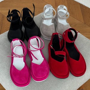 May include: An assortment of platform sandals in various colors. The selection includes black, gray, fuchsia, and red suede-like sandals. Each pair features an ankle strap with a buckle closure and a rounded toe design. The sandals are arranged on a neutral-toned surface.