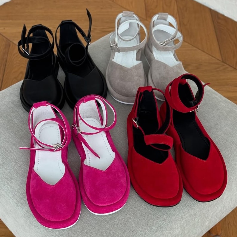 May include: An assortment of platform sandals in various colors. The selection includes black, gray, fuchsia, and red suede-like sandals. Each pair features an ankle strap with a buckle closure and a rounded toe design. The sandals are arranged on a neutral-toned surface.