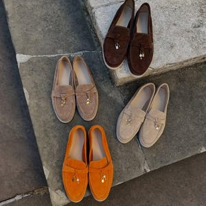 May include: Three pairs of suede loafers in dark brown, light tan, and bright orange. Each pair features a strap across the vamp adorned with a small metallic padlock and key charm. The shoes have a rounded toe and a flat, light-colored sole.