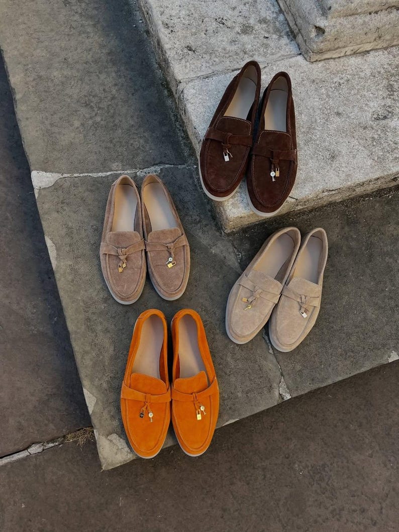 May include: Three pairs of suede loafers in dark brown, light tan, and bright orange. Each pair features a strap across the vamp adorned with a small metallic padlock and key charm. The shoes have a rounded toe and a flat, light-colored sole.