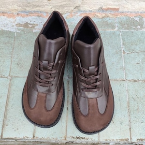 Può includere: Un paio di scarpe in pelle marrone con un design stringato. Le scarpe hanno una punta in pelle scamosciata marrone scuro e una tomaia in pelle liscia. Le suole sono nere e l'interno è foderato con un tessuto scuro. Le scarpe sono nuove.