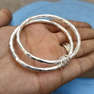 May include: Two silver-colored bangles with a hammered, textured finish. The two circular bracelets are linked together by a small section of thin silver-colored wire wrapped tightly around both bands, creating a single connected jewelry piece.
