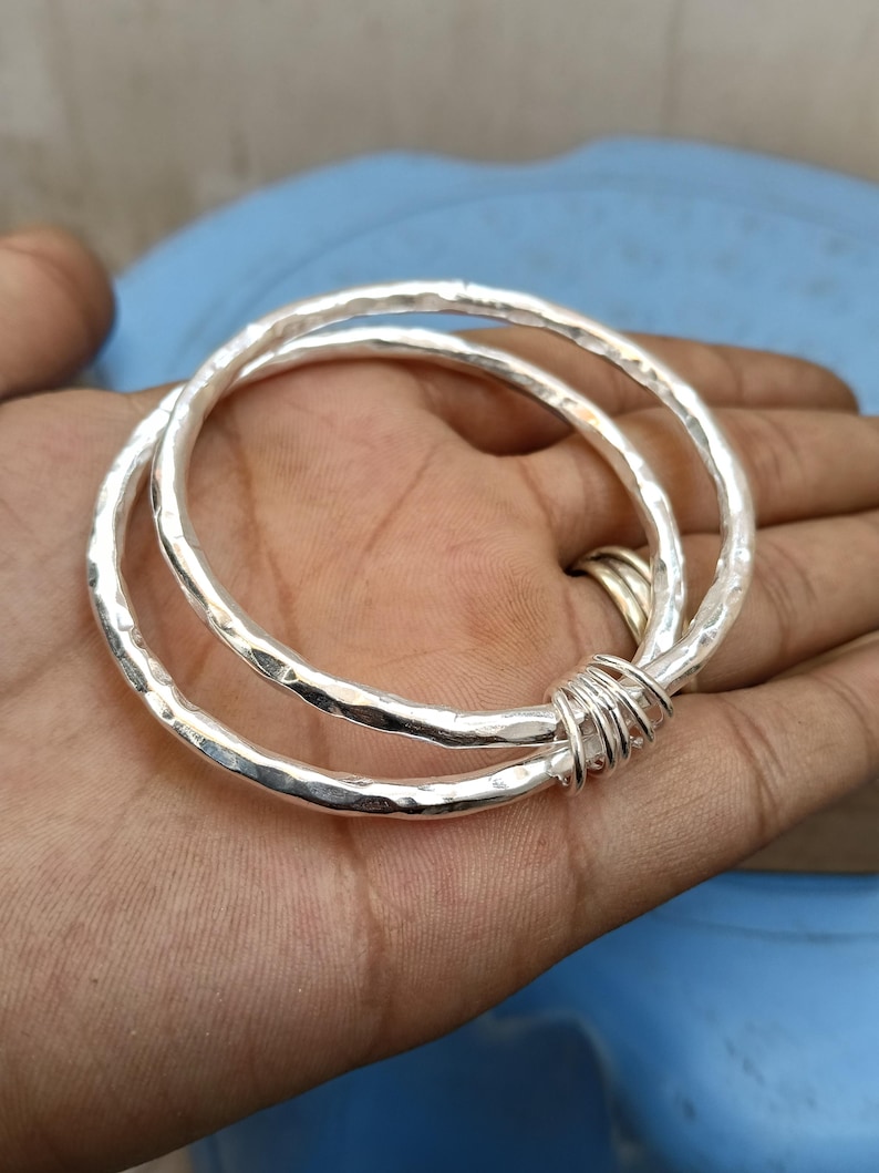 May include: Two silver-colored bangles with a hammered, textured finish. The two circular bracelets are linked together by a small section of thin silver-colored wire wrapped tightly around both bands, creating a single connected jewelry piece.