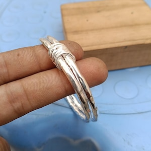 May include: A silver-colored bangle bracelet featuring a hammered, textured surface. The design consists of two parallel metal bands that are joined together by a series of tightly wrapped silver-colored wire coils at the top section.