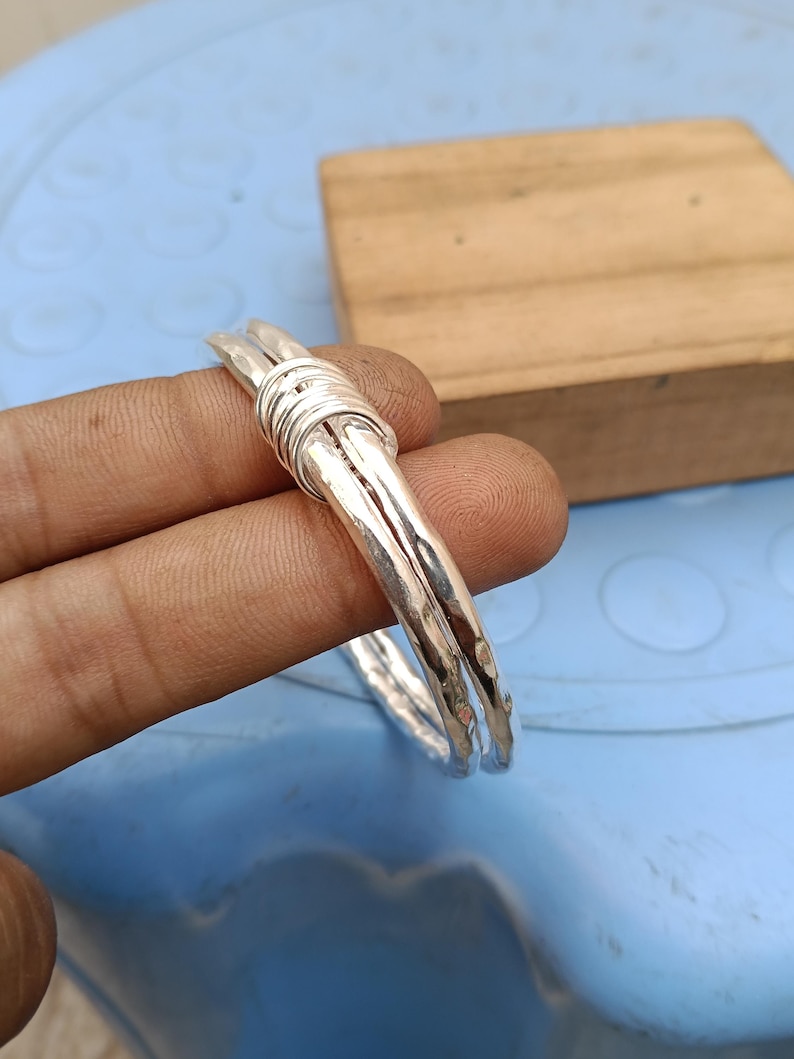 May include: A silver-colored bangle bracelet featuring a hammered, textured surface. The design consists of two parallel metal bands that are joined together by a series of tightly wrapped silver-colored wire coils at the top section.