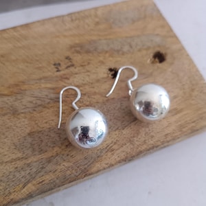 Puede incluir: Un par de pendientes de bola plateados con un diseño de gancho simple. Los pendientes son redondos y brillantes, reflejando la luz. Se exhiben sobre una superficie de madera.
