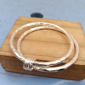 May include: A silver bangle bracelet featuring two interlocking circular bands with a hammered, textured finish. The two bands are joined together by a small, smooth, cylindrical silver connector piece that holds them in a fixed, overlapping position.